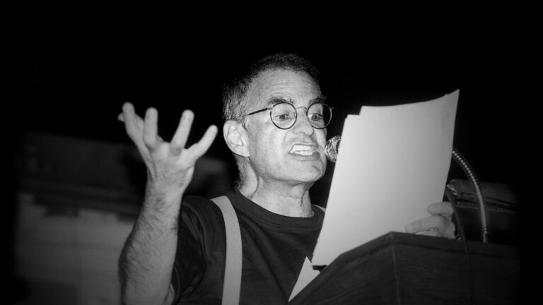 A black and white photo of Larry Kramer passionately speaking at a podium, holding a paper.