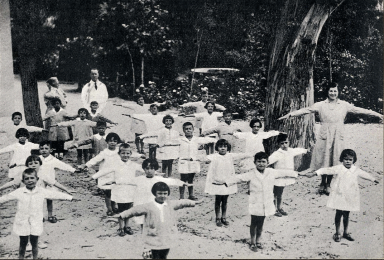 Children exercising outdoors with teachers standing with the arms out to their sides.