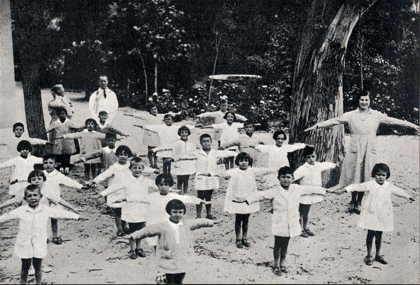 Children exercising outdoors with teachers standing with the arms out to their sides.