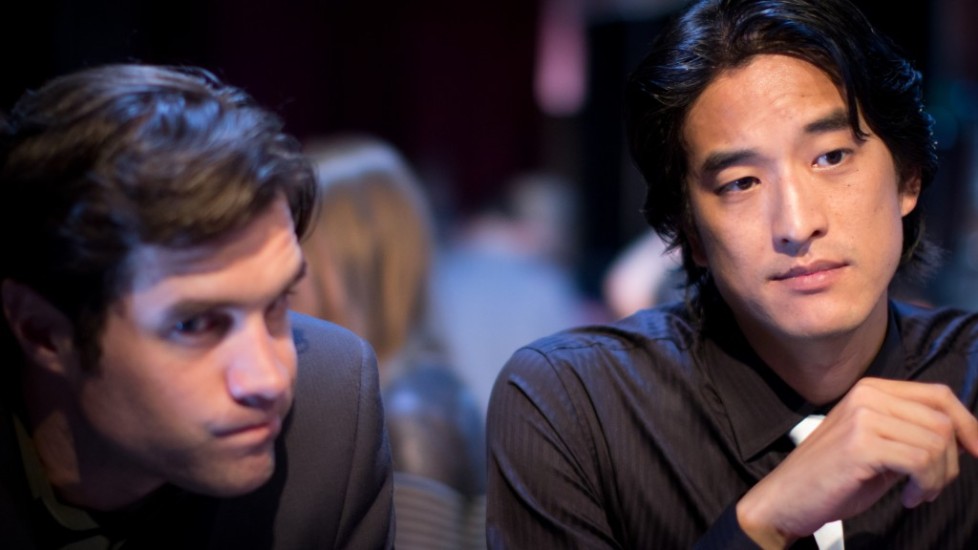Two men in suits, one Asian, in a dimly lit setting.