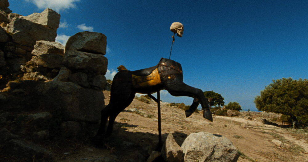 Surreal art: Skull atop a headless, antique horse sculpture near stone ruins against a clear blue sky.