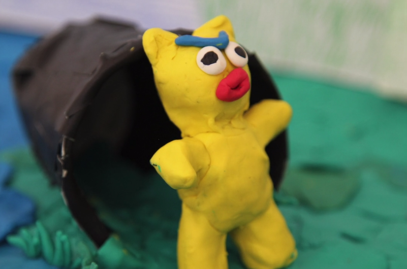Yellow clay figure emerging from a black clay barrel. Light House Studio Shorts art.