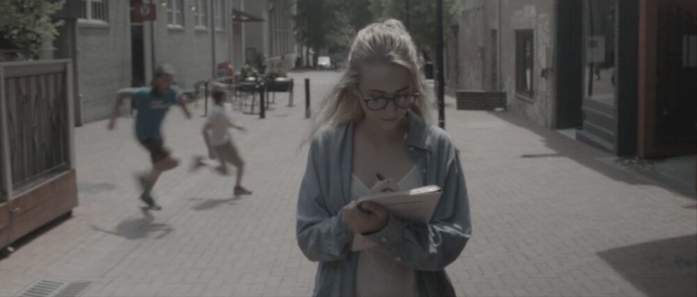 Woman with glasses writing in notebook, two boys running in background.