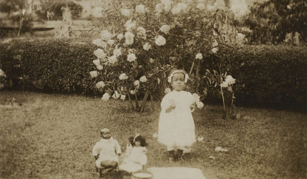 Vintage photo of a young girl in a white dress standing with her dolls in a garden with blooming flowers.