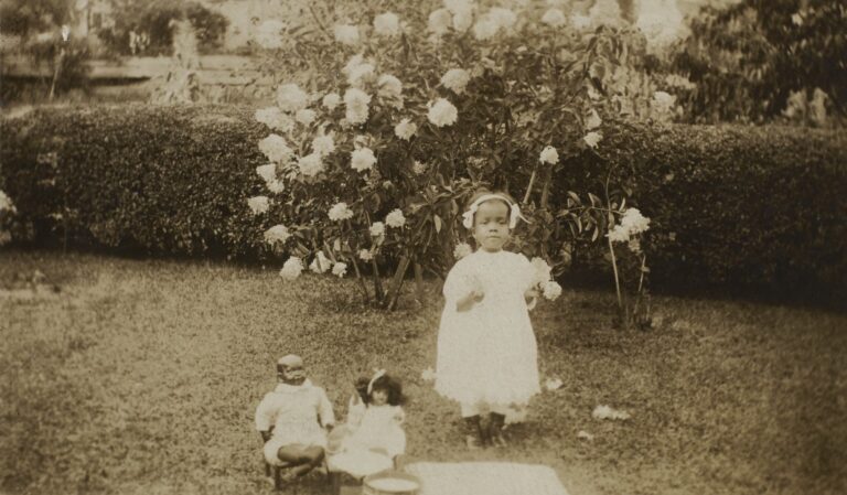 Vintage photo of a young girl in a white dress standing with her dolls in a garden with blooming flowers.
