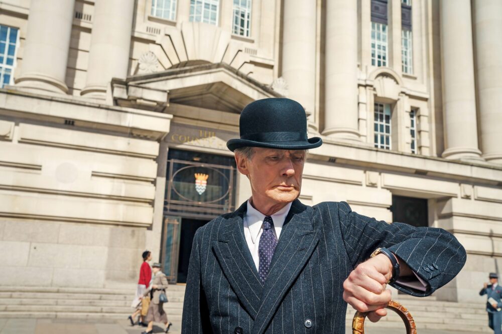 Man in bowler hat checking watch outside grand building.
