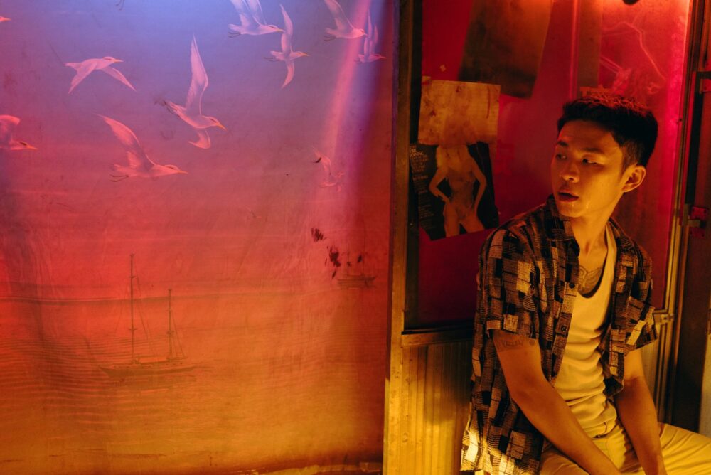 Asian man in patterned shirt, sits against a wall with a mural of seagulls and a sailboat.