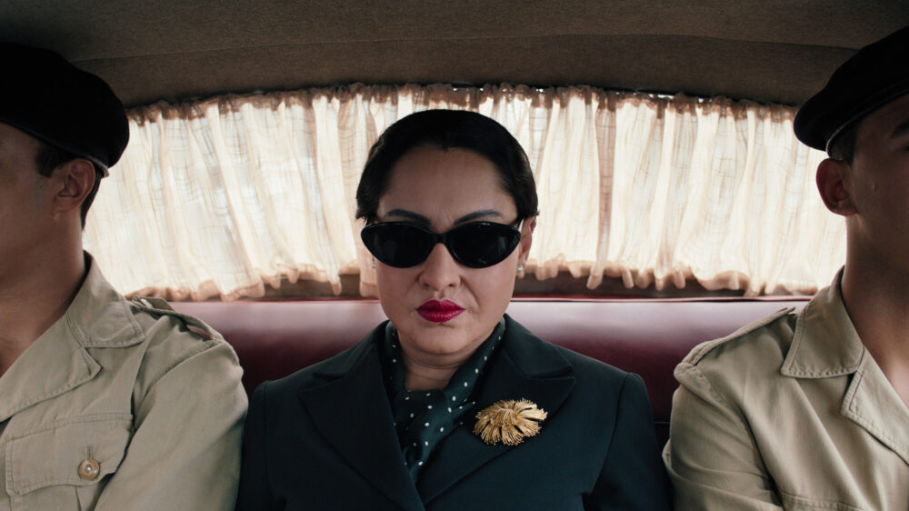 Oum Kulthum film still: Woman in sunglasses and a dark suit sits between two men in uniform.