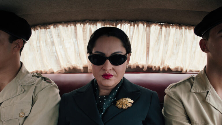 Oum Kulthum film still: Woman in sunglasses and a dark suit sits between two men in uniform.