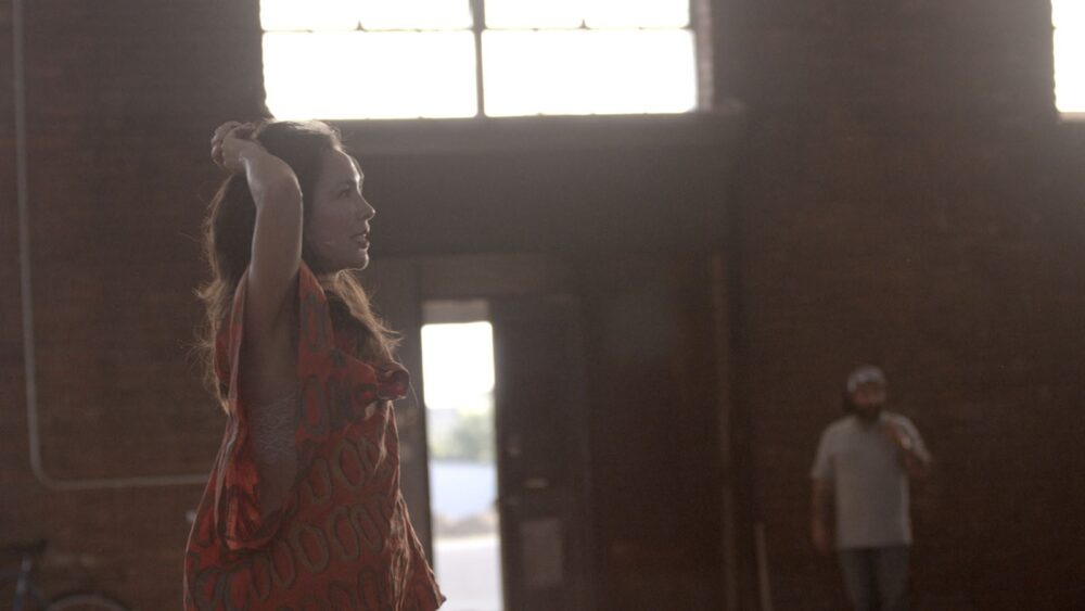 Woman in an orange top stands in a sunlit loft.