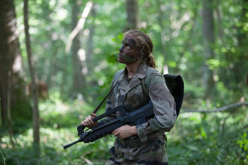 Female soldier with camouflage face paint holding a FAMAS rifle in a forest.