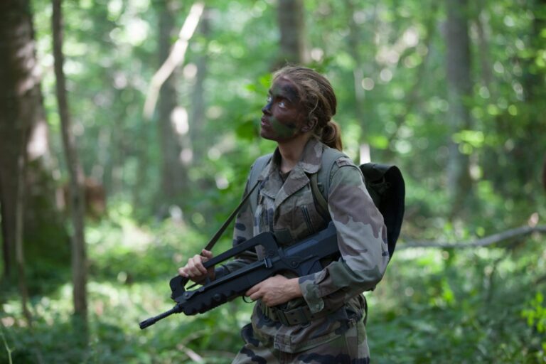 Female soldier with camouflage face paint holding a FAMAS rifle in a forest.