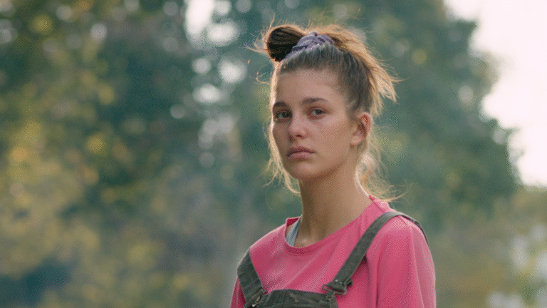 A young woman wearing overalls and a pink shirt with a messy bun.