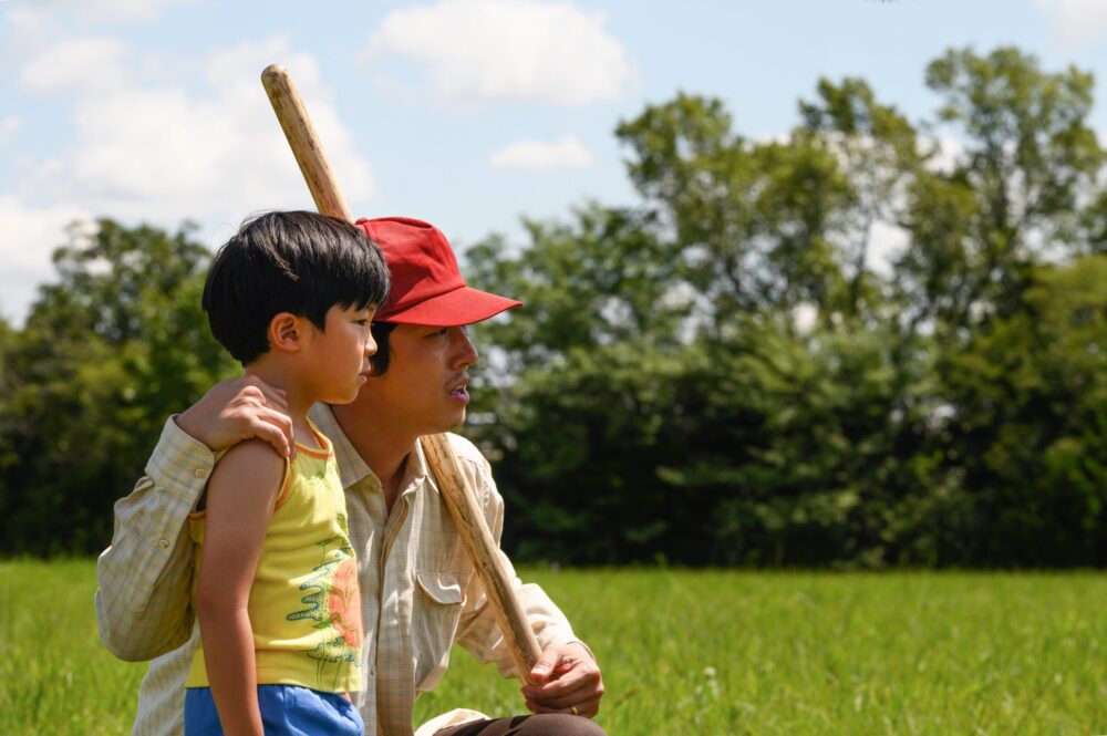 Father and son in Minari, kneeling in a field with a gardening tool, looking off into the distance.