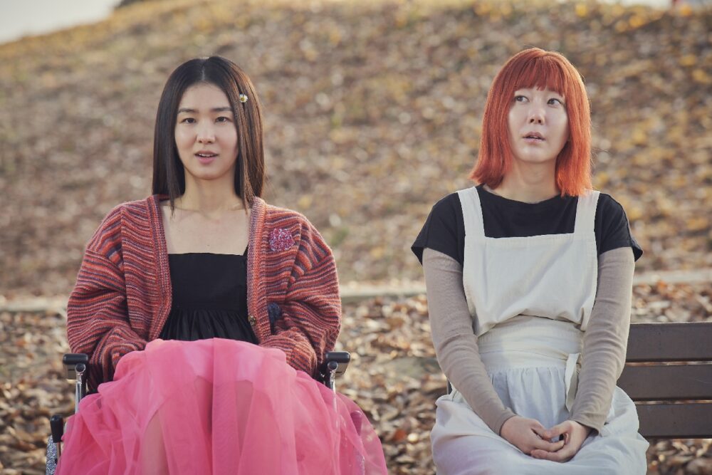 Two women, one in a wheelchair with a pink skirt, the other with red hair, sit outside. Ms. Apocalypse.
