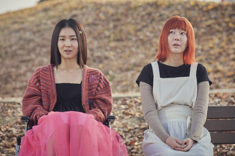 Two women, one in a wheelchair with a pink skirt, the other with red hair, sit outside. Ms. Apocalypse.