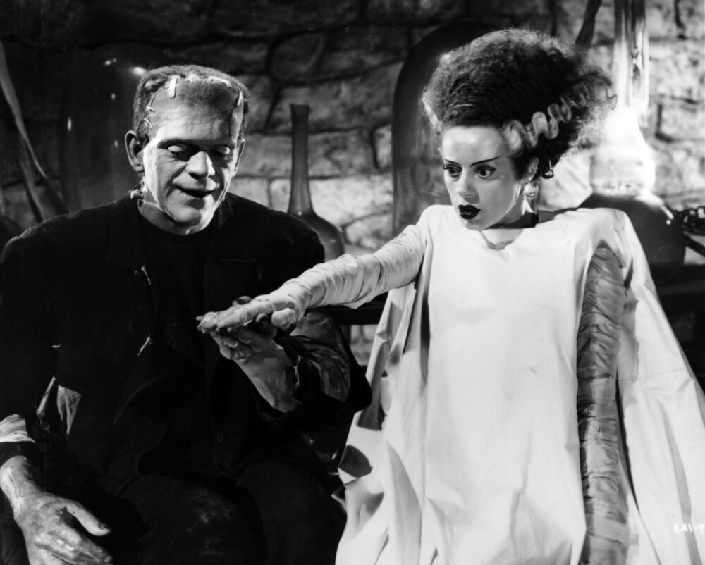 Bride of Frankenstein and Frankenstein's monster reaching out to each other in a classic horror film scene.