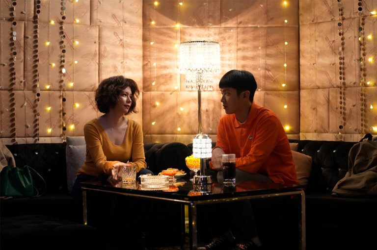 Woman and man talking at a table in a warmly lit bar. Mama Boy.
