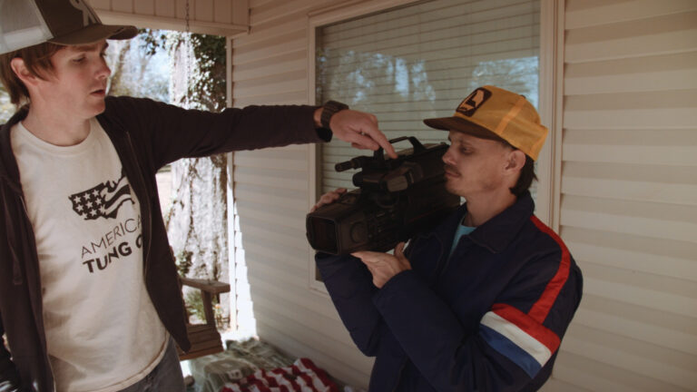 Man pointing at a vintage video camera held by another man in a Max Peril scene.