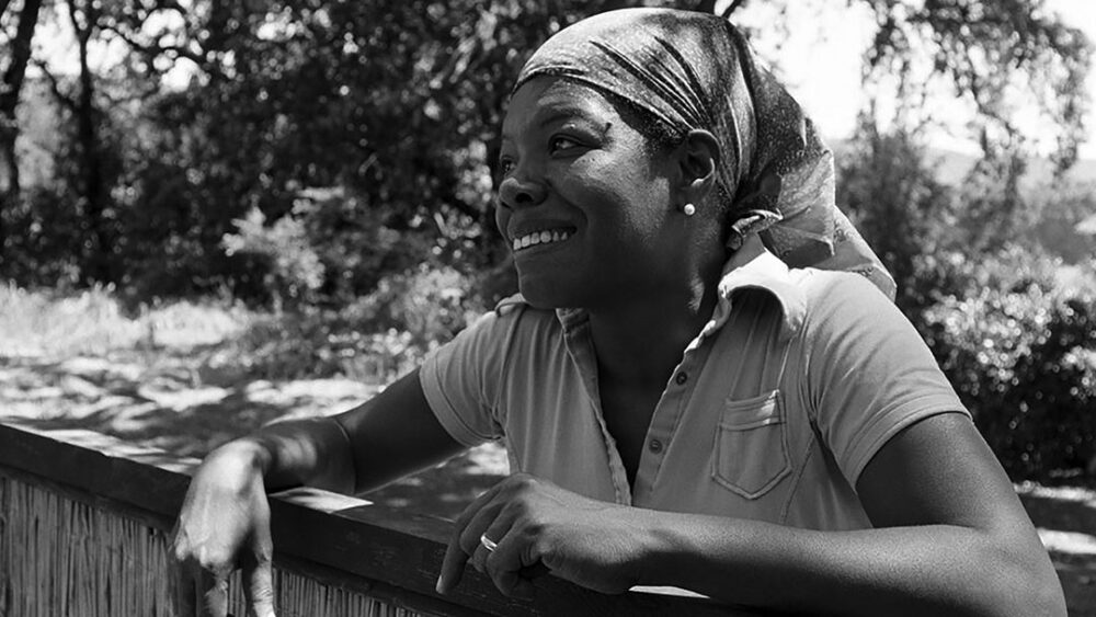Maya Angelou smiling, wearing a headscarf and leaning on a fence in a garden setting.