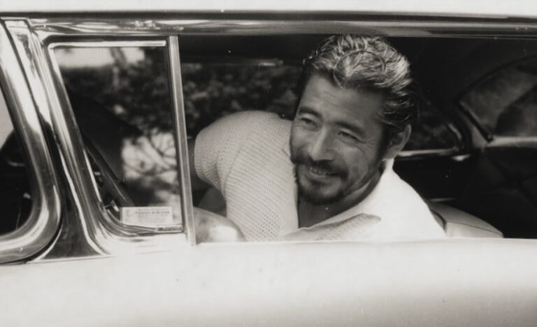 Toshiro Mifune, the Last Samurai, smiling from a car window, wearing a white shirt and light sweater.