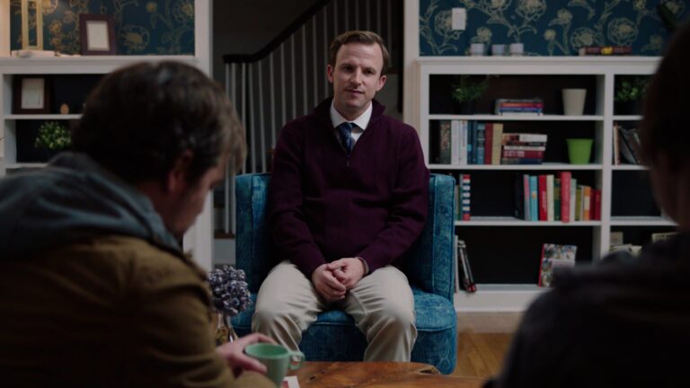 Man in maroon sweater sits in blue chair, facing two people in a therapy session.