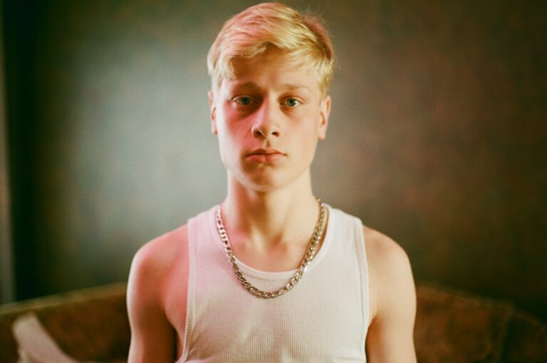 Young man with blonde hair wearing a white tank top and silver chain.