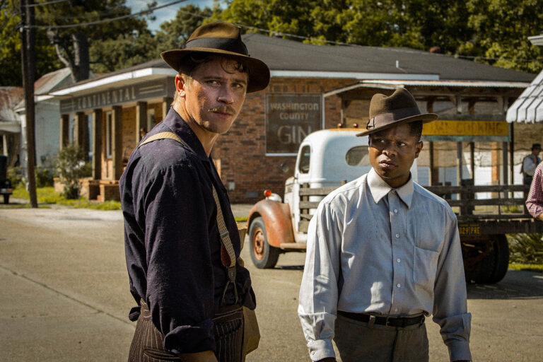 Two men in hats stand on a dusty road in "Mudbound." Vintage truck and building in background.