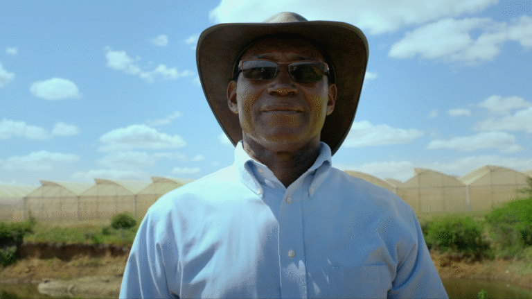 Mully in hat and sunglasses stands before greenhouses under a blue sky.