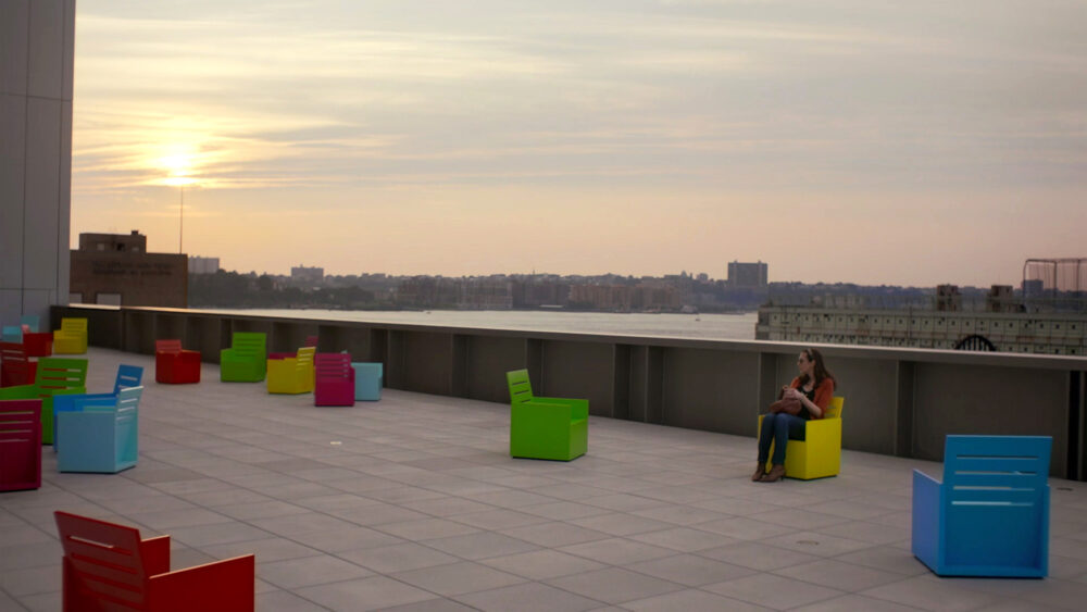 Woman sits in a yellow chair on a rooftop terrace with colorful chairs at sunset.