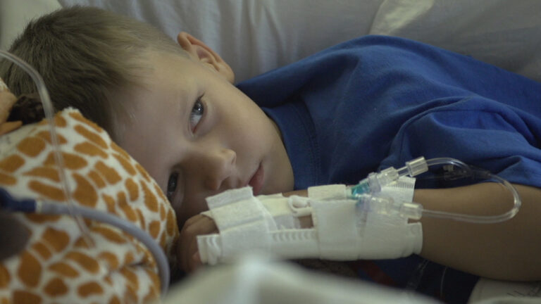 Child in hospital bed with IV, resting with stuffed giraffe.