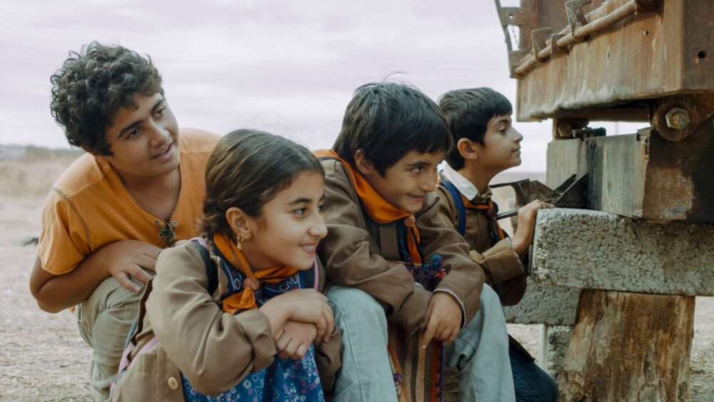 Children in "Neighbours" film peer at a structure, wearing scarves and jackets.