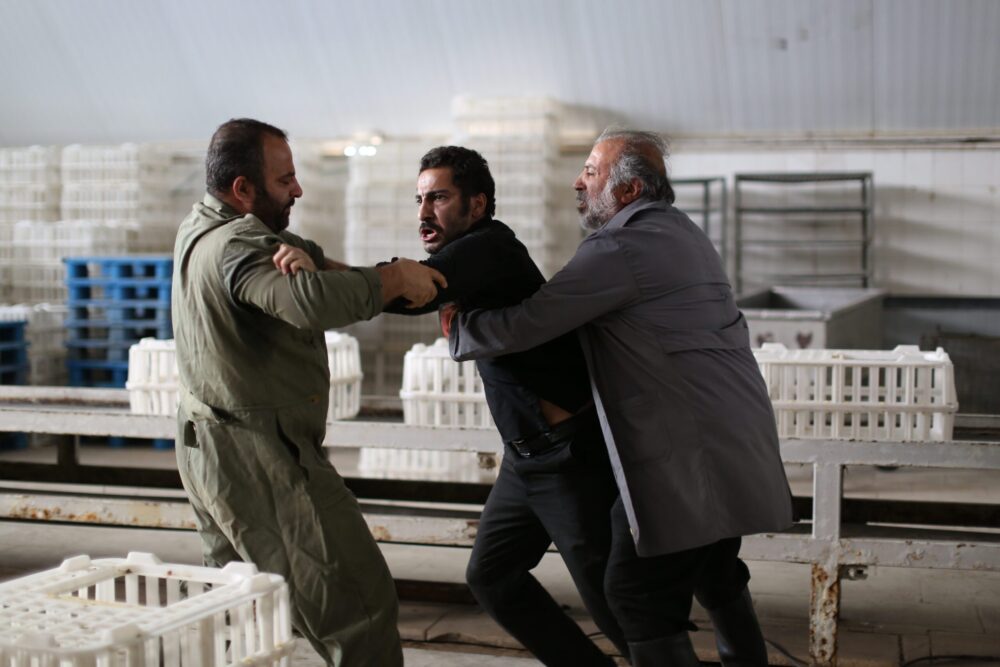 Scene from "No Date, No Signature" showing two men restraining a third in an industrial setting.