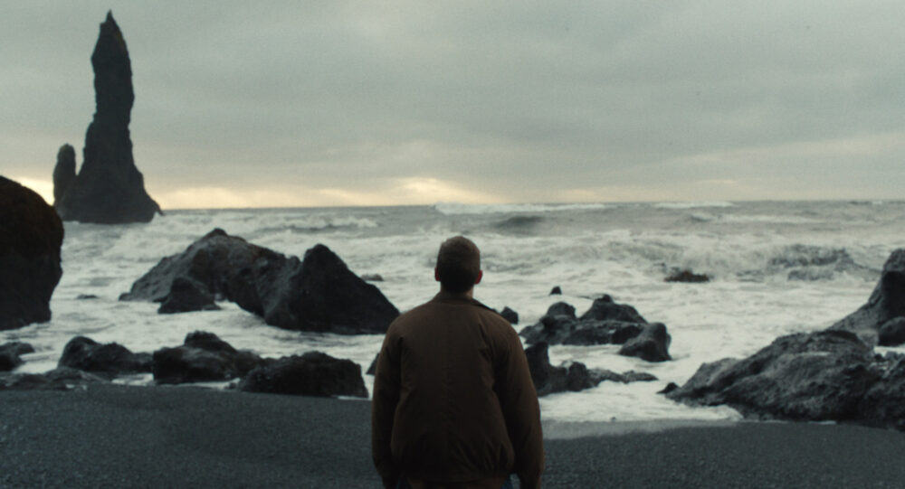 Man on black sand beach, possibly from "The Noise of Engines," gazing at sea stacks under cloudy sky.