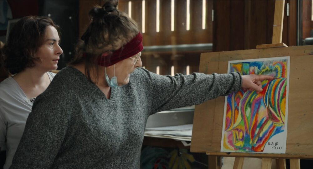 Two women discuss a vibrant, abstract painting on an easel. The painting is signed "A.A.B. 2021.