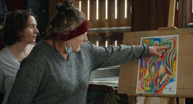 Two women discuss a vibrant, abstract painting on an easel. The painting is signed "A.A.B. 2021.