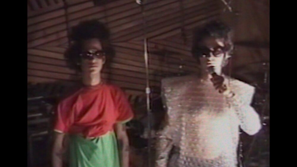 Omar and Cedric in a vintage, grainy still. Two figures in sunglasses and eccentric outfits.