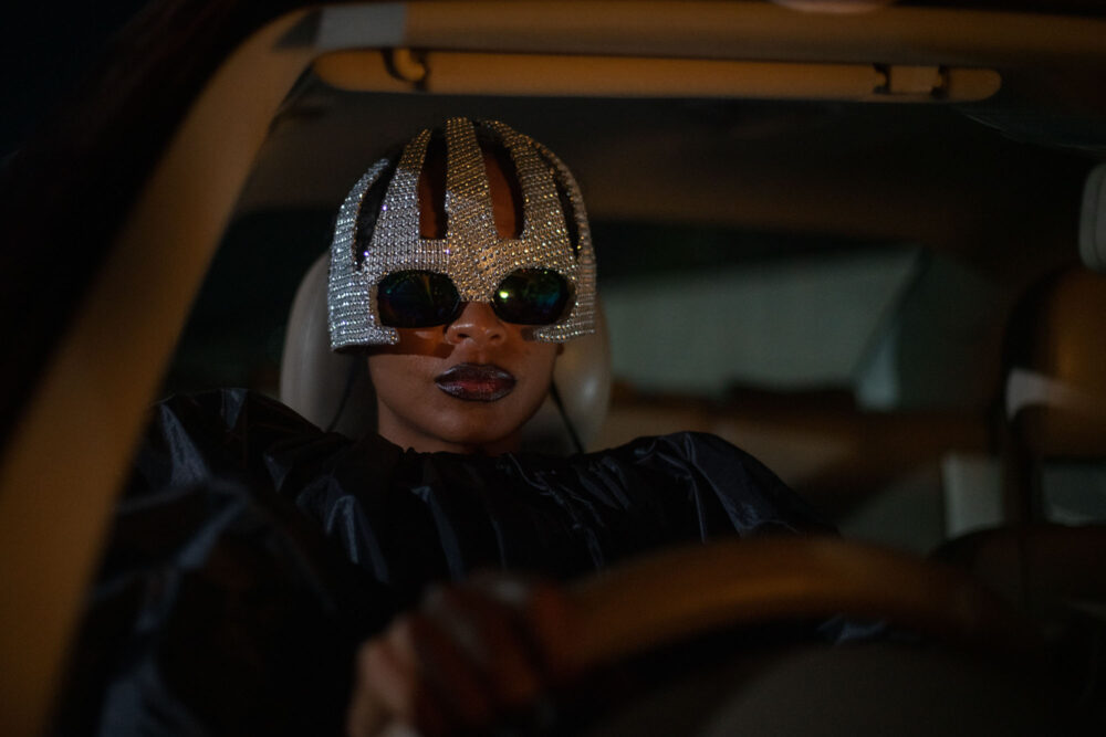Person in a car wearing a jeweled mask and sunglasses, driving at night.