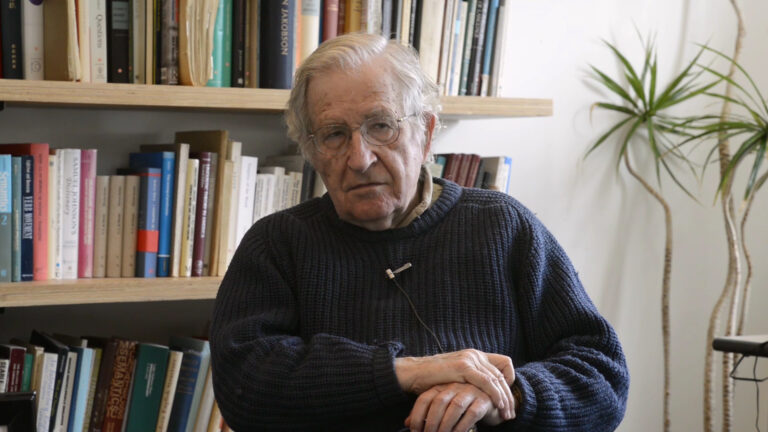 Noam Chomsky portrait with bookshelves, discussing Orwell's Revolution.