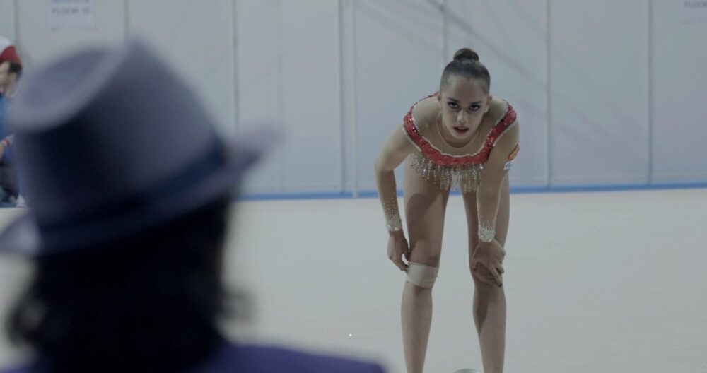 Rhythmic gymnast resting, leaning on knees, with coach in foreground.