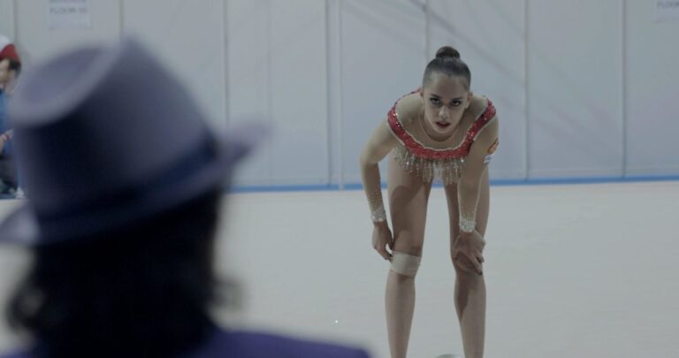 Rhythmic gymnast resting, leaning on knees, with coach in foreground.