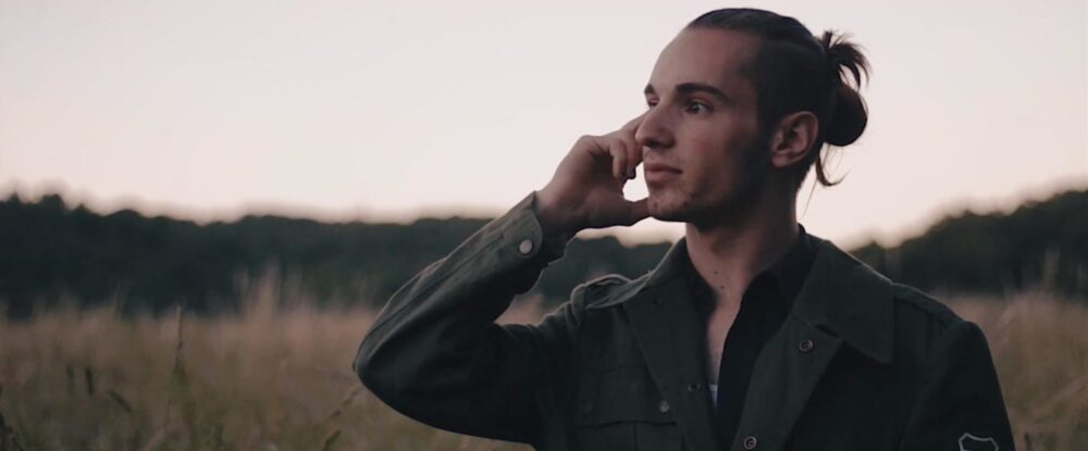 Man with a man bun in a field wearing an olive green jacket.