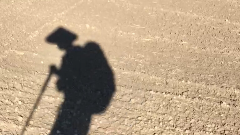Shadow of a pilgrim with hat, backpack, and walking stick on a dusty path.