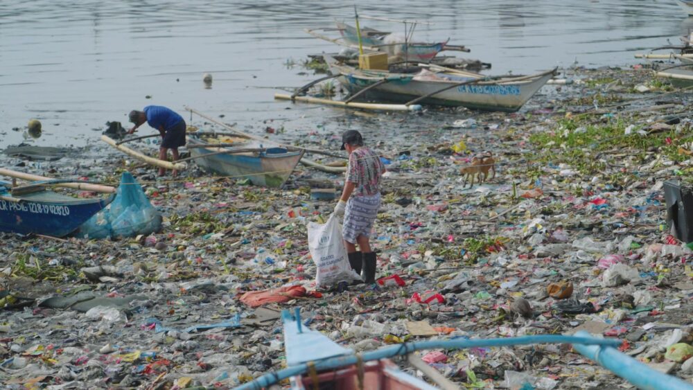 Man cleaning plastic waste from a polluted riverbank with boats; environmental issue.