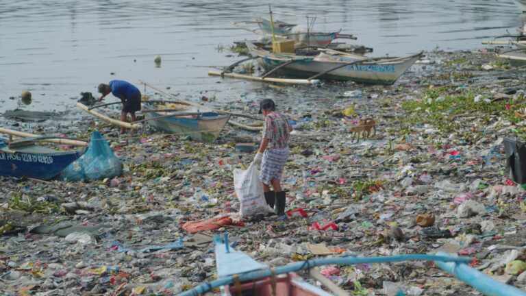 Man cleaning plastic waste from a polluted riverbank with boats; environmental issue.
