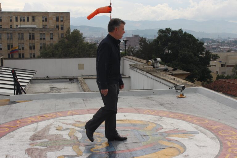 Former Colombian President Juan Manuel Santos walks on a helipad with the Colombian coat of arms, with a city view.