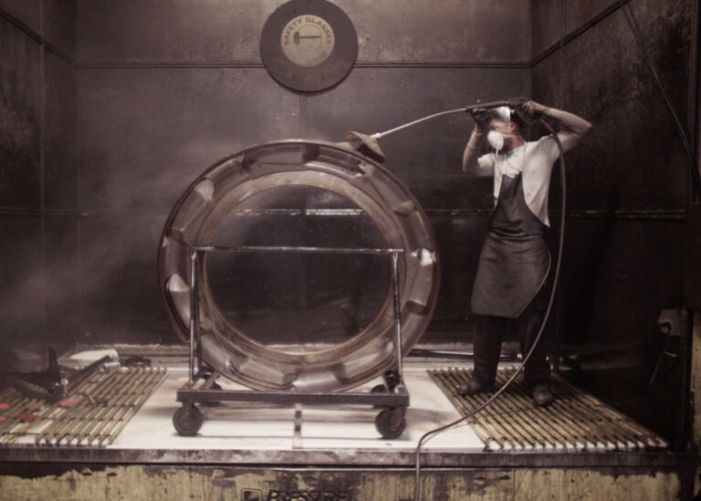 Worker using a pressure washer on a large metal ring, "Safety Glasses" sign visible.