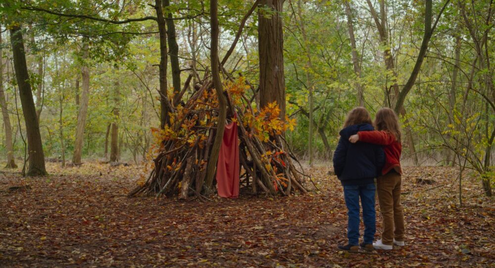 Two children stand before a stick hut in the woods. Petite Maman film scene.