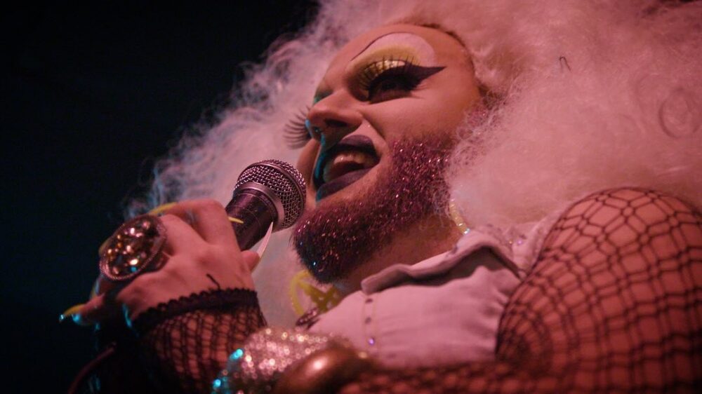 Drag queen with glitter beard singing into a microphone, wearing fishnet sleeves and a large ring.