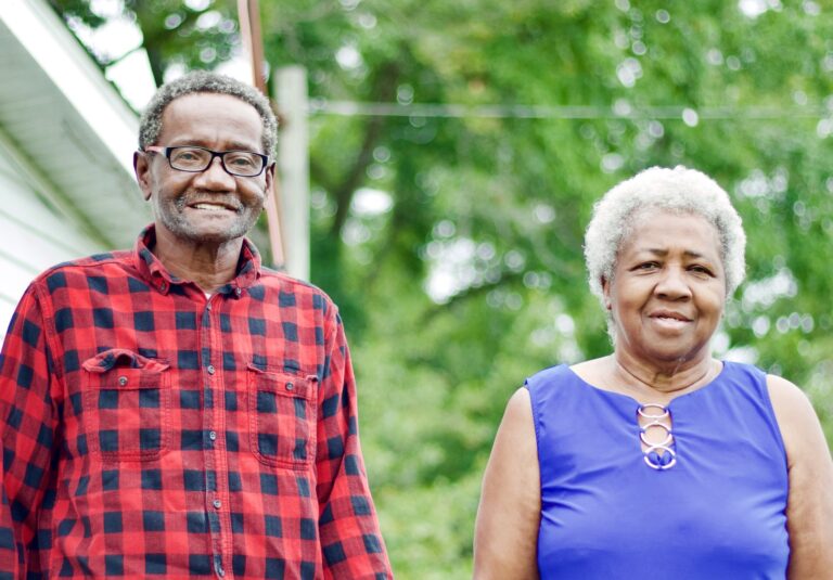 Elderly African American couple smiling outdoors; man in red flannel, woman in blue top.